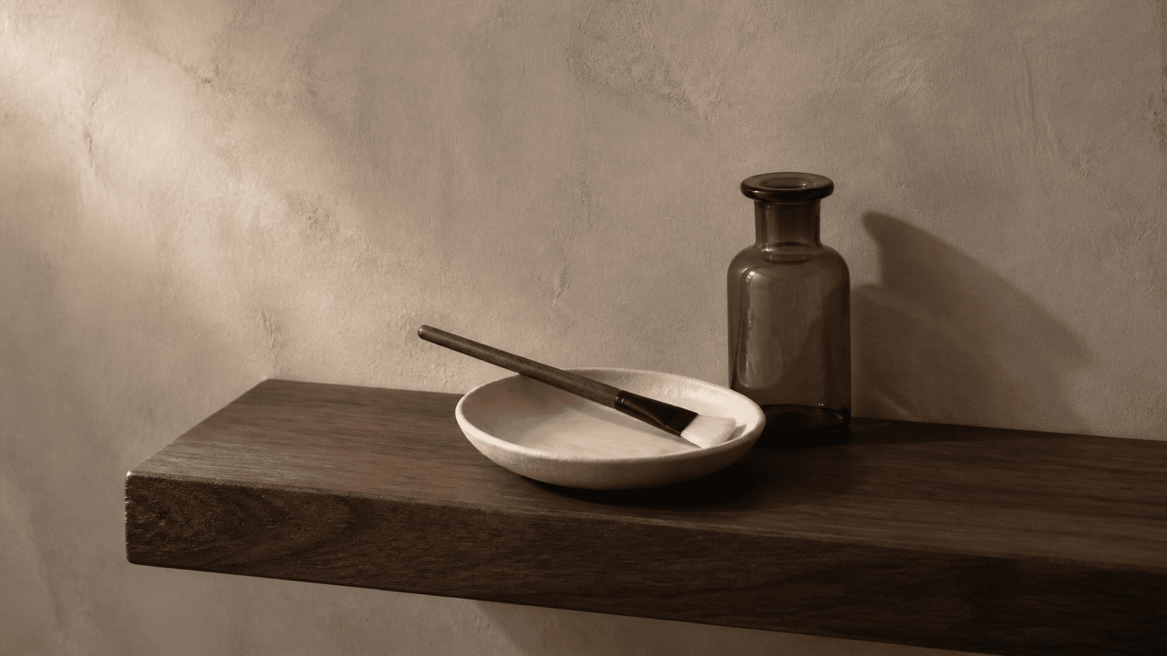 A facial brush rests across an ivory ceramic dish beside an unlabeled smoked-glass bottle on a walnut shelf