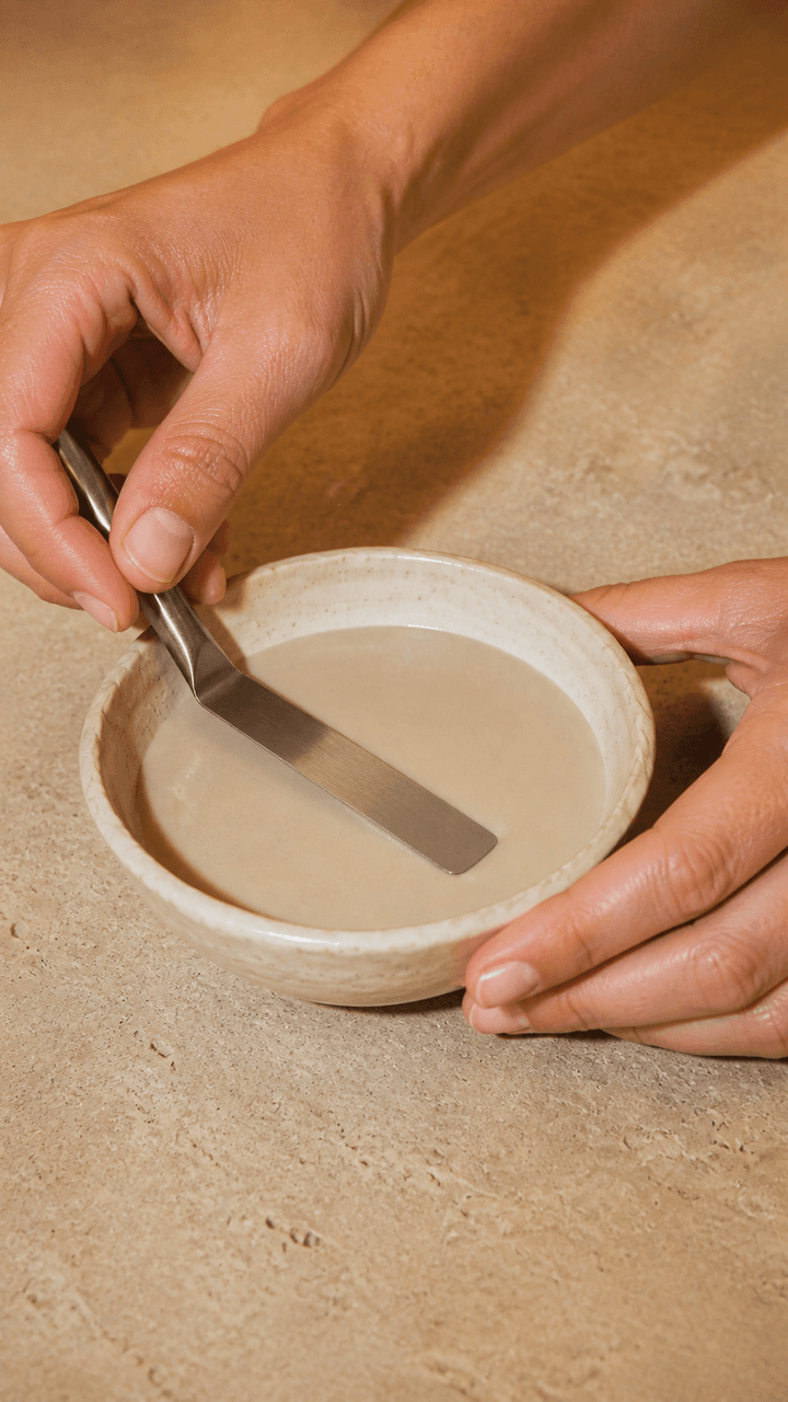 Two hands hold a ceramic bowl and flat spatula while mixing a pale masque